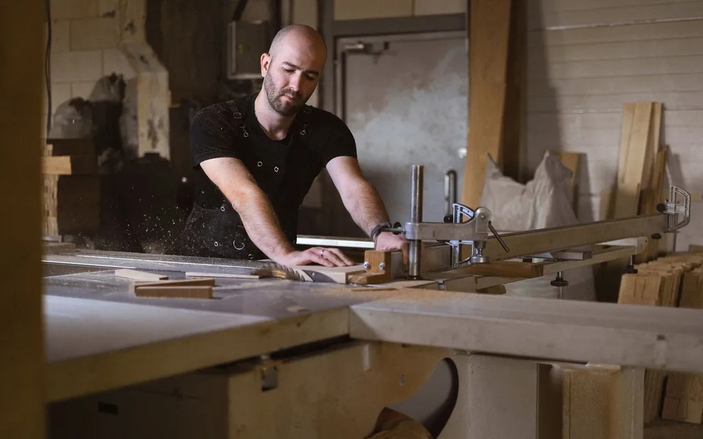 Carpenter in an apron guiding a timber board through a table saw in a workshop