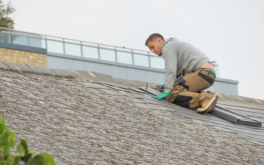 Roofer kneeling on a pitched roof hammering a slate into place